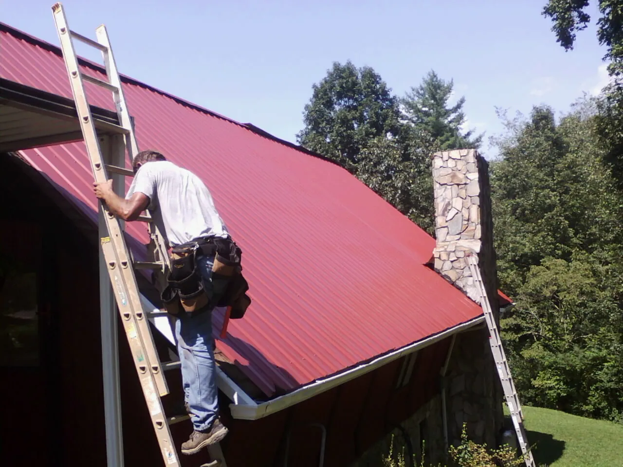 Roofer on a ladder beside a red metal roof and stone chimney — Soffit & Fascia Repair by Roofing Brothers in Rocky Ridge